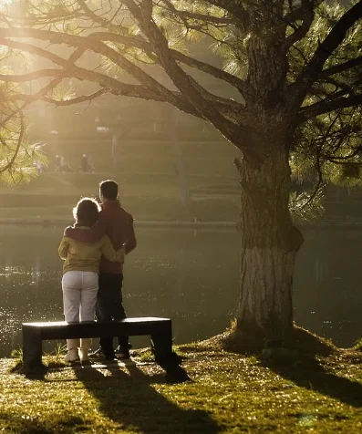 An older couple standing arm in arm by a lake, under a tree in evening sunlight